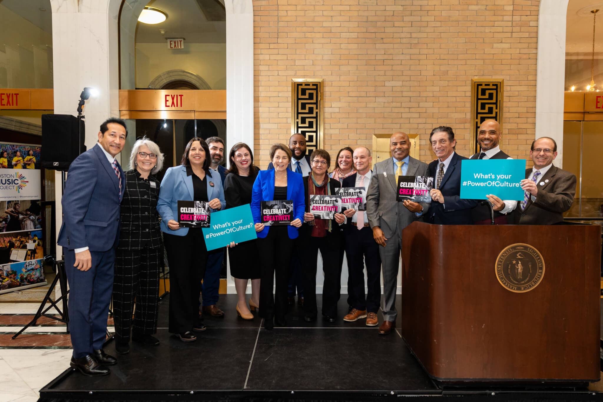 Fourteen people pose together in the State House holding creative advocacy signs.